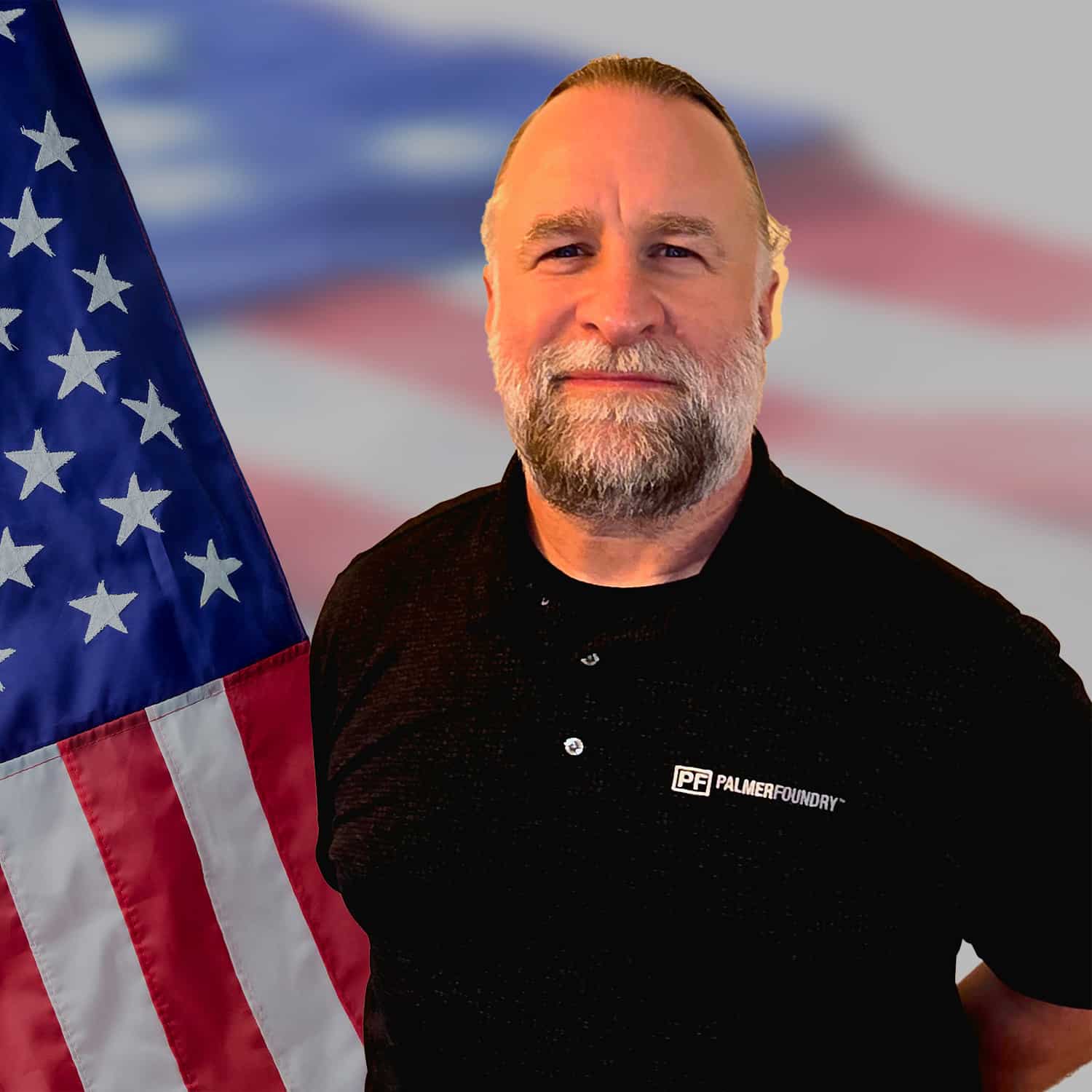 A man with a beard wearing a black Palmer Foundry shirt stands beside an American flag, with a blurred flag background.