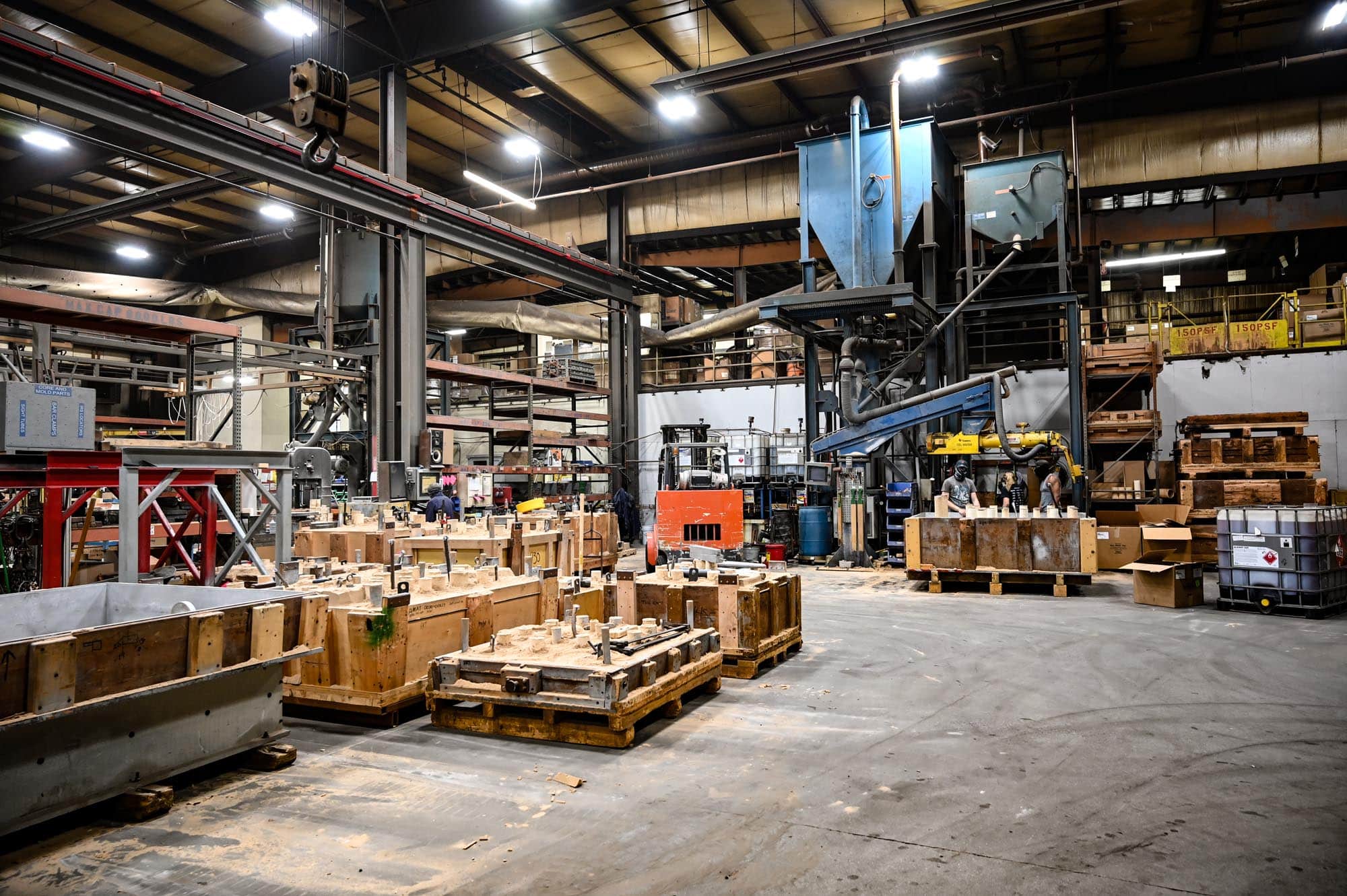 A large, busy industrial factory interior with machinery, wooden crates, and equipment scattered around; workers are visible in the background, and the space is lit by overhead lights.