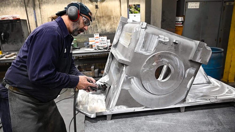 A man wearing safety gear uses a power tool to work on a large metal machine part in an industrial workshop in east windsor, ct