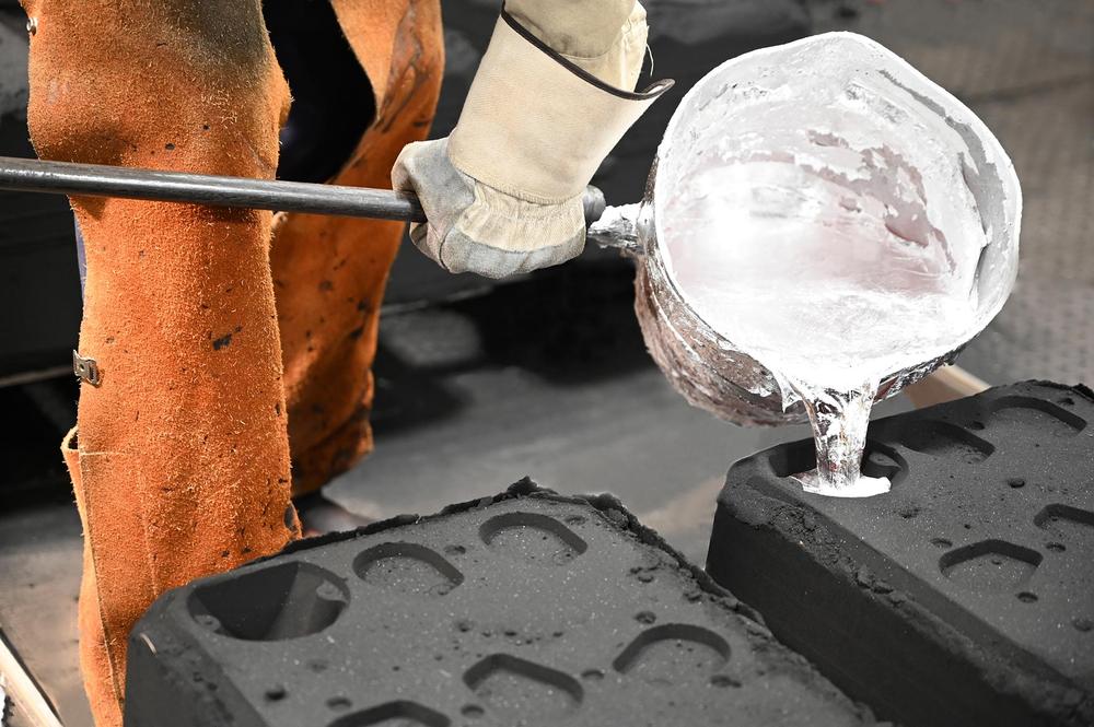 A worker in protective gloves and clothing pours molten metal from a crucible into a black mold during a metal casting process in an industrial setting in Suffield, CT